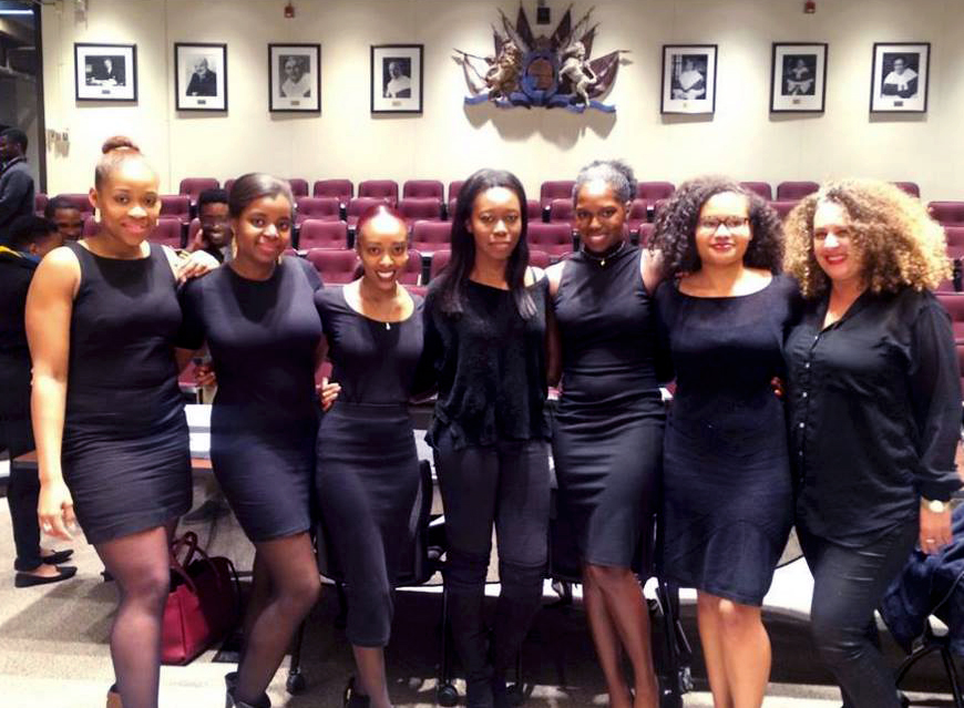 Seven panelists wearing black dresses standing with their arms around each other.